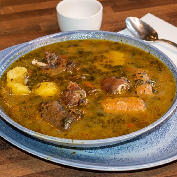 A blue bowl of Paolo's Caribbean Fusion soup with meat, potatoes, and vegetables on a wooden table.